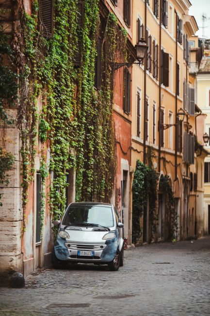 A narrow street in Trastevere, Rome, paved with wet cobblestones, with outdoor seating for cafés consisting of several wooden tables covered by large cream umbrellas, surrounded by green and wooden c